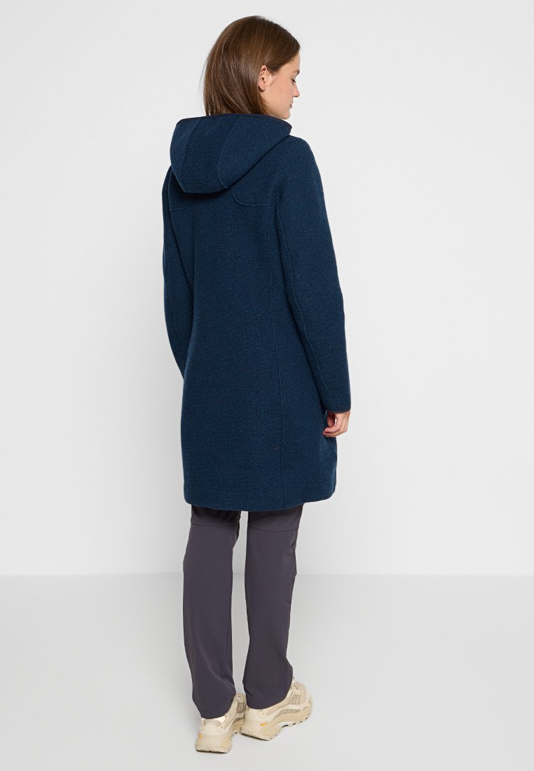 Cappotto di lana blu navy con cappuccio, caratterizzato da un tessuto strutturato, maniche lunghe e lunghezza fino al ginocchio. Indossato sopra pantaloni scuri e scarpe beige.