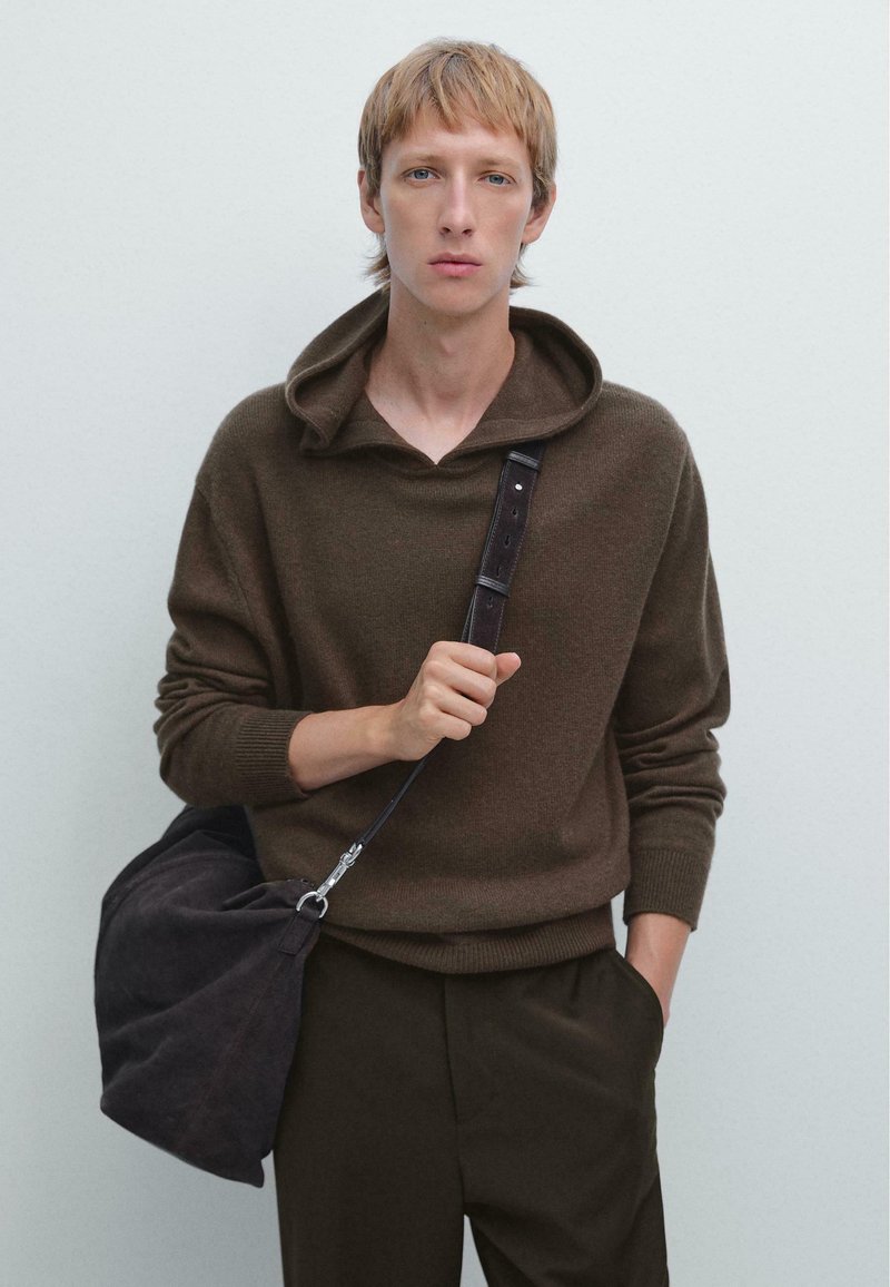 Bruine gebreide hoodie met een ruime halslijn en lange mouwen. Gecombineerd met donkere broeken en een zachte, donkere schoudertas. Soepele textuur.