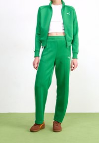 Veste de survêtement verte avec des bandes blanches sur les côtés et une fermeture éclair, associée à un pantalon de survêtement vert. Présente un design ajusté et un haut blanc en dessous. Baskets marron.
