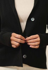 Czarny kardigan wełniany z długimi rękawami, z wykładanym kołnierzem i szarymi guzikami. Modelka zapina guziki nad kremową bluzką.