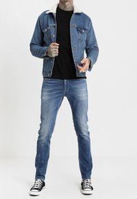 Denim takki, jossa on shearling-niska, siniset farkut ja musta t-paita. Asusteina mustat lenkkarit valkoisilla pohjilla ja tatuoinnit käsivarsissa.