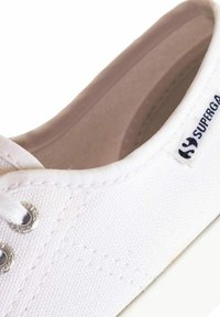 Sneaker bianca in tela con occhielli argentati e lacci bianchi. Presenta una texture liscia e un'etichetta di marca sul lato. Design semplice e a profilo basso.