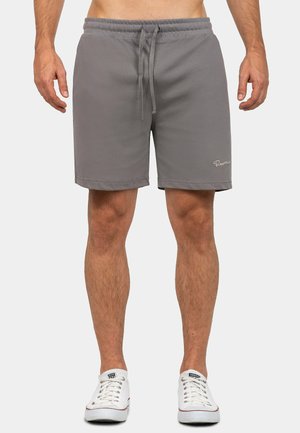 Man draagt grijze shorts met trekkoord en witte lage sneakers, staand tegen een effen lichte achtergrond.