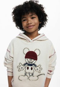 Niño sonriente con cabello rizado que lleva un hoodie color crema con un ratón de dibujos animados que tiene un gorro de lana rojo y detalles en rojo cosidos.