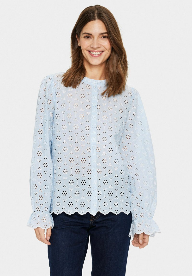 Blusa azzurra chiara con bottoni, caratterizzata da un motivo floreale ricamato, orlo scallop e maniche lunghe con polsini arricciati. Indossata con jeans scuri.