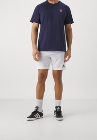 Námořnická modrá bavlněná tričko s krátkým rukávem a malým logem na hrudi, v kombinaci s bílými šortkami a černými teniskami Adidas.