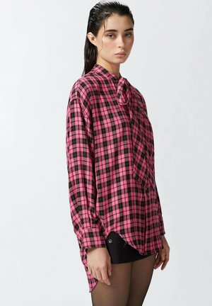 Camisa de cuadros rosa y negra con mangas largas, cuello y detalle de lazo en la parte frontal. Presenta un corte holgado y un dobladillo asimétrico. Textura de tela suave.