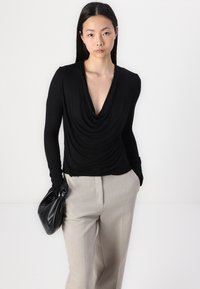 Long-sleeve black top with a draped cowl neckline, paired with beige trousers, held with a black leather bag. Smooth, lightweight fabric.