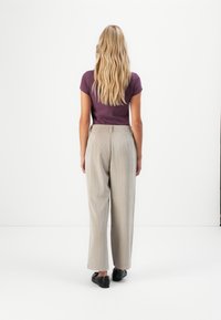 Frau mit langen welligen blonden Haaren, die ein lila Hemd, beige weite Hosen und schwarze Schuhe trägt, steht mit dem Rücken zu einem weißen Hintergrund.