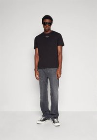 Vīrietis valkā melnu beanie cepuri, lielas melnas saulesbrilles, melnu T-kreklu ar logo, izbalējušas pelēkas džinsus un melnus zema profila sporta apavus ar baltiem zoliem.