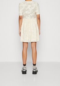 Kurzärmliges, cremefarbenes Kleid mit strukturiertem Blumenmuster, geraffter Taille und schwingendem Rock. Kombiniert mit klobigen schwarzen Schuhen und gemusterten Socken.