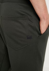 Dark green jogger pants made of smooth fabric, featuring a back pocket and a small black label. The design includes an elastic waistband.