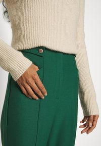 Personne portant un pull crème à côtes rentré dans un pantalon vert taille haute avec une poche boutonnée, une main reposant sur la poche.