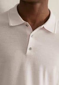 Helles beige Polohemd mit weicher Textur, ausgestattet mit einem Kragen, drei Knöpfen und einer glatten Stoffoberfläche. Minimalistisches Design mit zurückhaltenden Details.