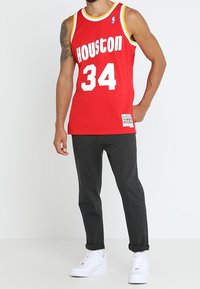 Mann trägt ein rotes Basketball-Trikot von Houston mit der Nummer 34, dunkelgraue Hosen, weiße Sneakers und ein Tattoo am linken Arm. Er steht mit der Hand in der Tasche.