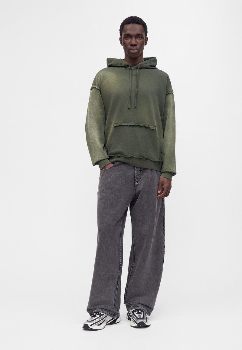 Homme debout portant un sweat à capuche vert olive avec des bords effilochés, un jean gris ample et des baskets blanches et noires sur un fond uni.