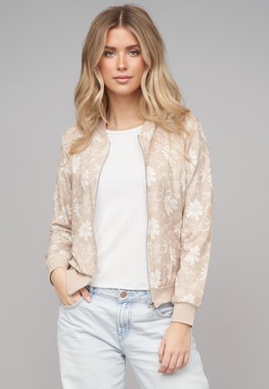 Giovane donna con capelli biondi indossa una giacca bomber beige a fiori, maglietta bianca e jeans azzurri, in piedi con una mano in tasca.