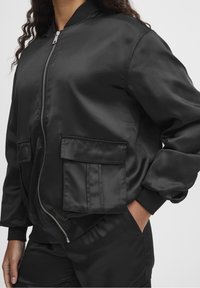 Schwarze Satin-Bomberjacke mit Seitentaschen, einem Vorderreißverschluss und gerippten Bündchen. Glatte Textur und lockere Passform.