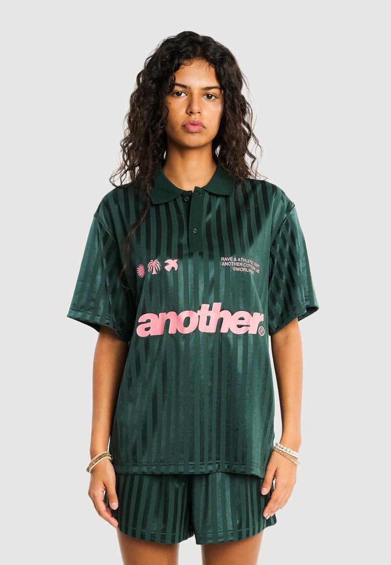 Femme aux cheveux bouclés portant un polo rayé vert foncé et un short assorti, avec le texte rose « another » sur le devant du polo.