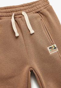 Brune bomulds-sweatpants med elastisk talje, hvide snore, sidelommer og et "Enjoy and Explore"-mærke i farverig tekst.