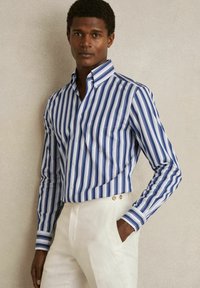 Chemise boutonnée rayée bleue et blanche avec col et manches longues, associée à un pantalon de couleur claire. Comprend une poche poitrine.