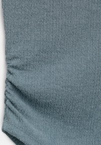 Gros plan d'un tissu texturé bleu clair avec des coutures froncées sur le côté gauche, montrant des détails de surface doux et irréguliers.