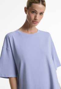 Světle fialové oversized tričko, krátké rukávy, kulatý výstřih, hladká bavlněná tkanina, volný střih, viditelné švy na rukávech.