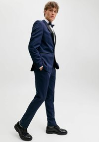 Giovane uomo che cammina con sicurezza indossando uno smoking blu navy con scarpe eleganti nere e un papillon, su uno sfondo bianco semplice.