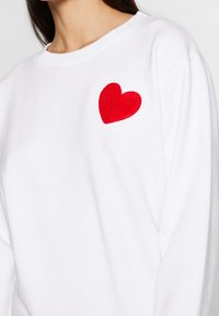 Bílý svetr vyrobený z bavlny s červenou plsťovou aplikací ve tvaru srdce na levém prsou. Má ribovaný crew neck a dlouhé rukávy.