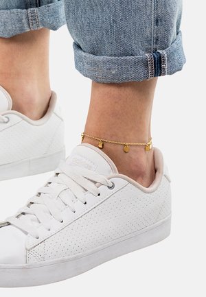 Hvite perforerte sneakers med flate lisser og en beige aksent. Ankelarmbåndet består av en gullkjede med små runde vedheng. Rullet denimcuffs.