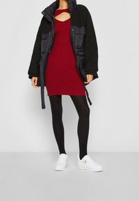 Rotes geripptes Kleid, geschichtet mit einer schwarzen gepolsterten Jacke mit plüschiger Textur. Schwarze Strumpfhosen und weiße Sneakers vervollständigen den Look.