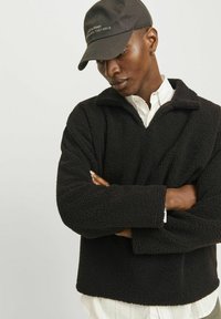 Schwarzer Fleece-Pullover mit hohem Kragen und strukturiertem Oberflächenmaterial, kombiniert mit einer schwarzen Mütze mit gesticktem Schriftzug.