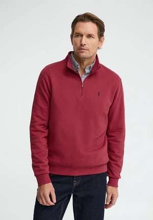 Homme portant un pull rouge à demi-fermeture éclair sur une chemise rayée à col, associé à un jean bleu foncé, se tenant devant un fond clair uni.