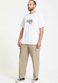 Camiseta blanca de algodón con un estampado gráfico negro, combinada con pantalones cargo beige que tienen bolsillos laterales y zapatillas negras.