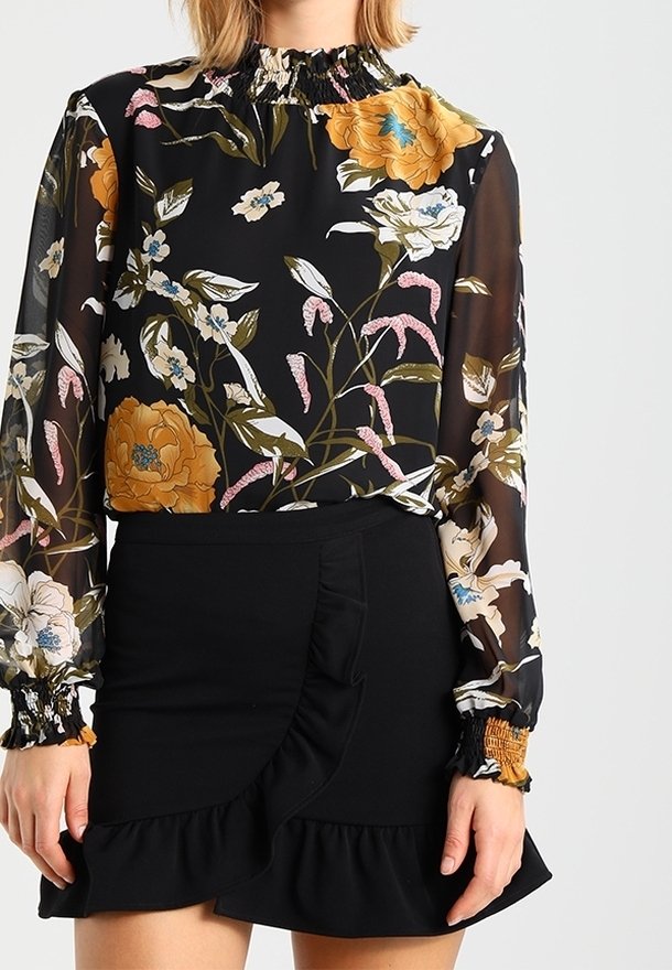 Femme portant un chemisier noir transparent à motifs floraux jaunes, blancs et roses, assorti à une mini-jupe noire à volants.
