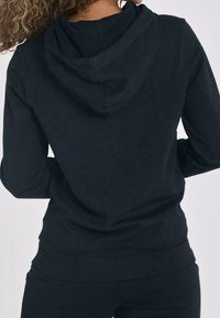 Felpa nera con cappuccio regolabile, maniche lunghe e taglio aderente. Il tessuto sembra morbido e liscio, adatto per un abbigliamento casual.
