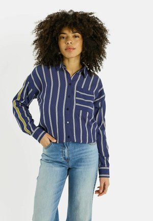 Mujer con cabello rizado, vestida con una camisa azul con botones y rayas blancas, y jeans azul claro, posando con una mano en el bolsillo.