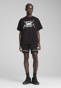 T-shirt en coton noir avec un texte graphique blanc, associé à un short noir avec des rayures grises. Le modèle porte des baskets noires et des chaussettes.