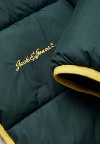 Mørkegrøn oppustelig jakke med gule detaljer. Har glat stof, horisontale quiltede sektioner og broderet "Jack & Jones" logo.