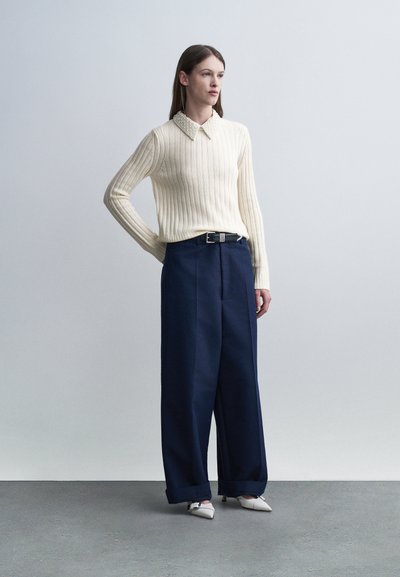 Femme debout portant un pull crème côtelé avec un col orné, un pantalon large bleu marine avec ceinture, et des chaussures blanches à talons pointus.