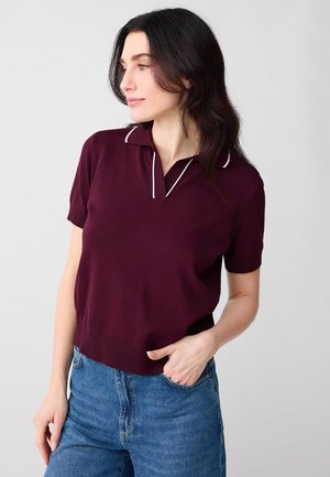 Femme aux cheveux foncés portant un polo bordeaux à manches courtes avec un liseré blanc et un jean bleu, debout avec une main dans la poche.
