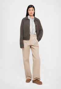 Cardigan marrone a maglia con colletto rotondo e bottoni frontali, abbinato a pantaloni beige con ricami floreali e scarpe con platform marroni.