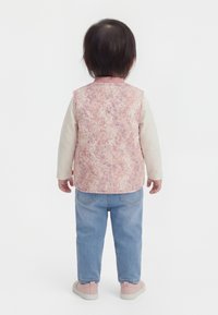 Taapero seisoo selinpäin, pukeutuneena vaaleanpunaiseen kukkakuosiseen liiviin, kermaan väriseen pitkähihaiseen paitaan, vaaleansinisiin farkkuihin ja vaaleanpunaisiin kenkiin.