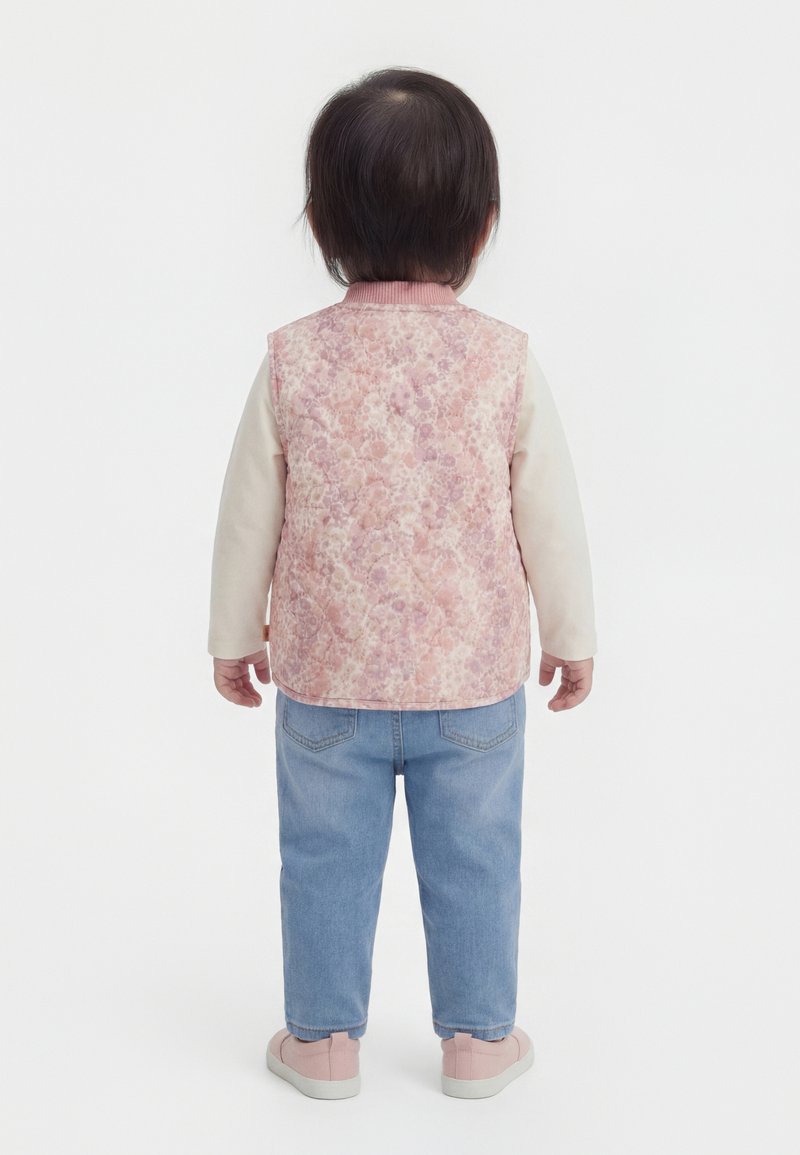 Taapero seisoo selinpäin, pukeutuneena vaaleanpunaiseen kukkakuosiseen liiviin, kermaan väriseen pitkähihaiseen paitaan, vaaleansinisiin farkkuihin ja vaaleanpunaisiin kenkiin.