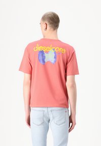 Kortermet, korallfarget t-skjorte med grafik på baksiden, som har teksten "dieselcore" i gult og et abstrakt blått formdesign. Laget av bomull.