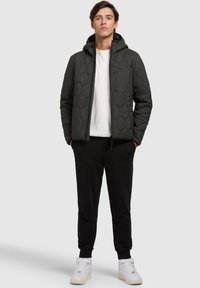 Quilted green jacket with a hood, featuring hexagonal patterns, front zip pocket, worn over a white shirt, paired with black pants and white shoes.