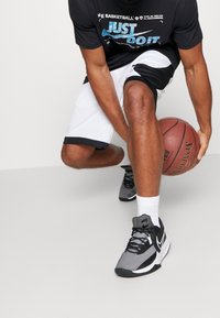 Zapatillas deportivas grises y negras con parte superior de malla, suelas blancas y detalles de cordones. La persona viste pantalones cortos negros y una camiseta mientras sostiene un balón de baloncesto.