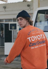 Oranžová mikina s potlačou "Toyota Land Cruiser", so zaokrúhleným výstrihom a voľným strihom. Nosí sa s tmavou čiapkou, ktorá má štruktúrovanú tkaninu.