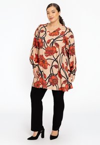 Blomstret bluse med røde blomster på en beige baggrund; har lange ærmer og en V-halsudskæring, parret med sorte slim-fit bukser og højhælede sko.