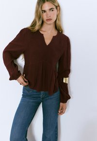 Blouse peplum bordeaux avec un col en V, tissu texturé, et manches longues, présentant un accent doré sur le bras gauche. Associée à un jeans bleu.